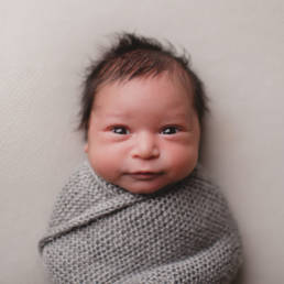 awake newborn baby swaddled in a grey knit blanket stares into camera