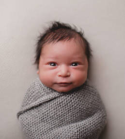 awake newborn baby swaddled in a grey knit blanket stares into camera