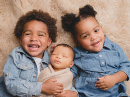 pre school aged older brother and sister hold swaddled newborn baby in between them, posed on back looking up at camera